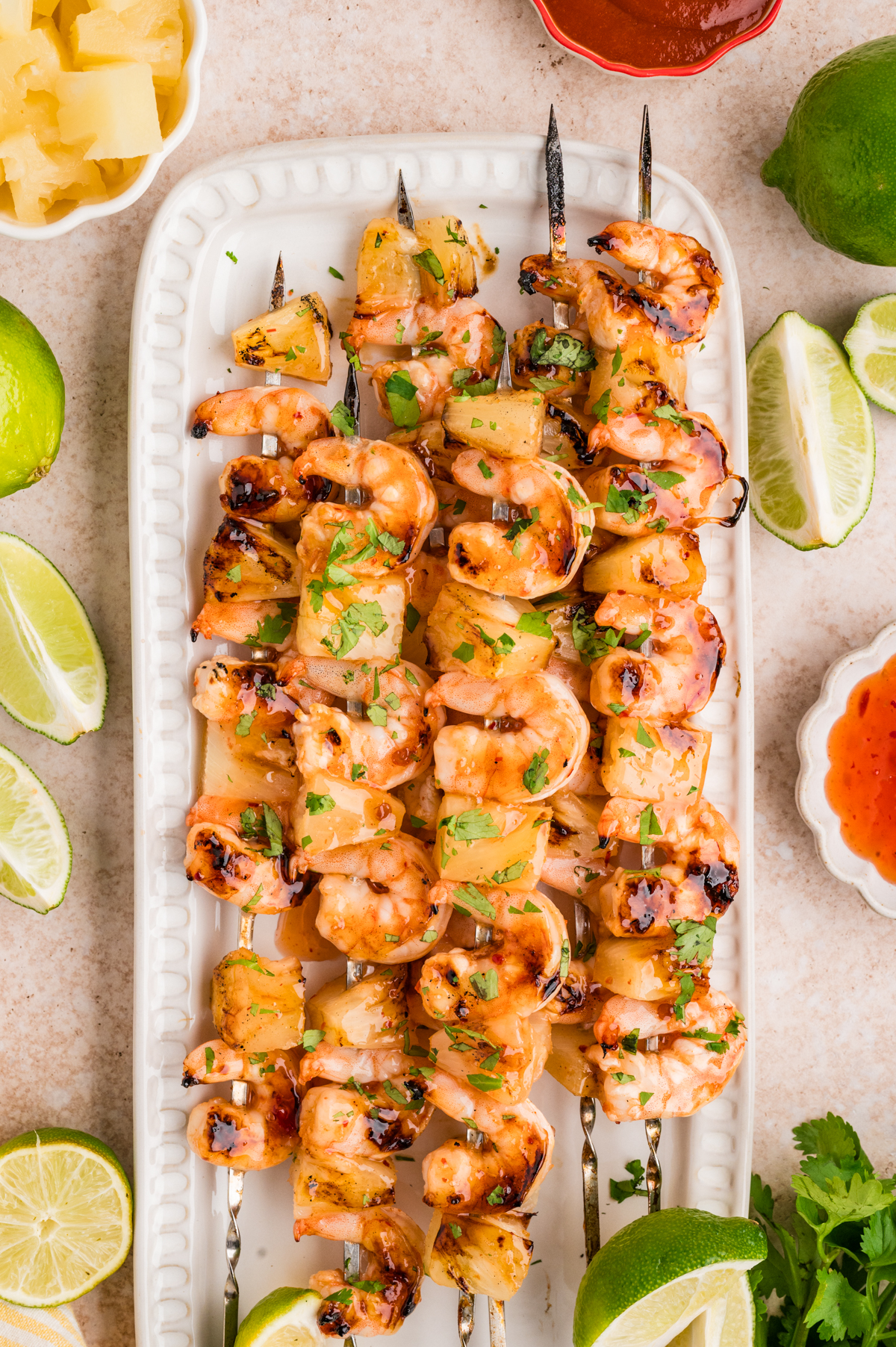 Shrimp Pineapple Skewers are served grilled on a white rectangular plate, garnished with chopped herbs and surrounded by lime wedges and small bowls of sauces.