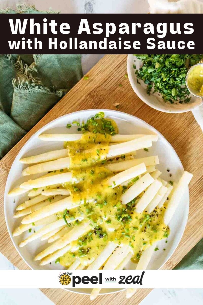 Plate of white asparagus spears topped with hollandaise sauce and chopped herbs on a wooden board, with extra herbs and sauce in small bowls nearby.