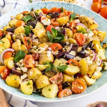 A bowl of potato salad with cherry tomatoes, black olives, red onion, feta cheese, and fresh herbs.