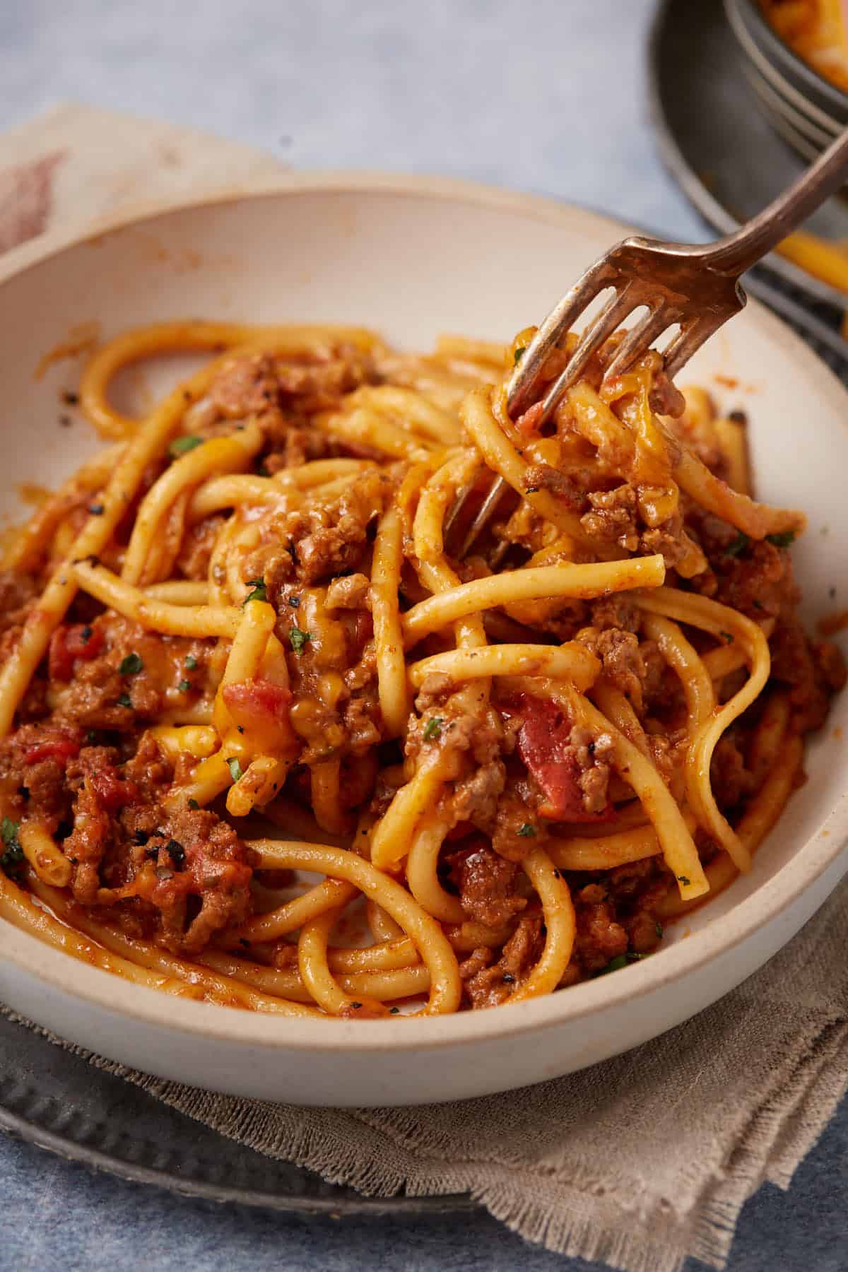 A bowl of bucatini pasta with meat sauce, garnished with herbs, being twirled with a fork.