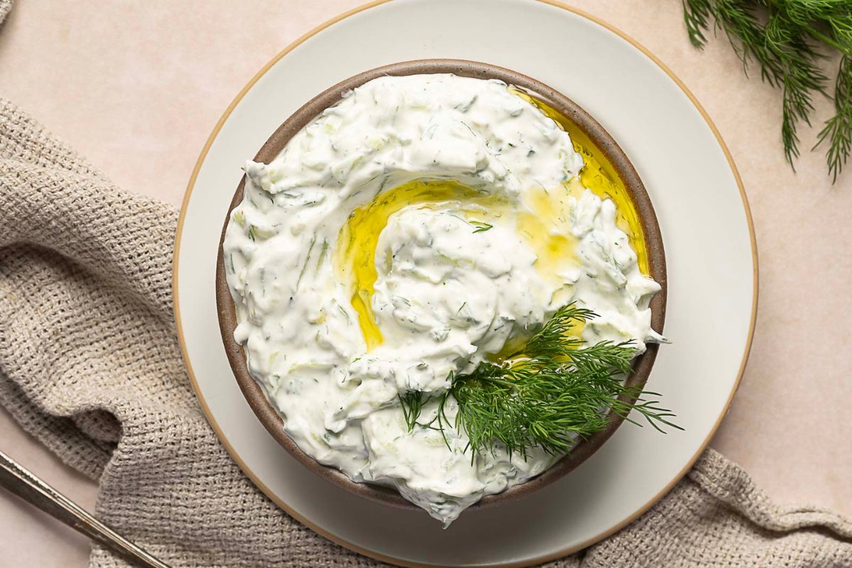 A bowl of tzatziki sauce garnished with fresh dill and a drizzle of olive oil, placed on a plate with a textured cloth nearby.
