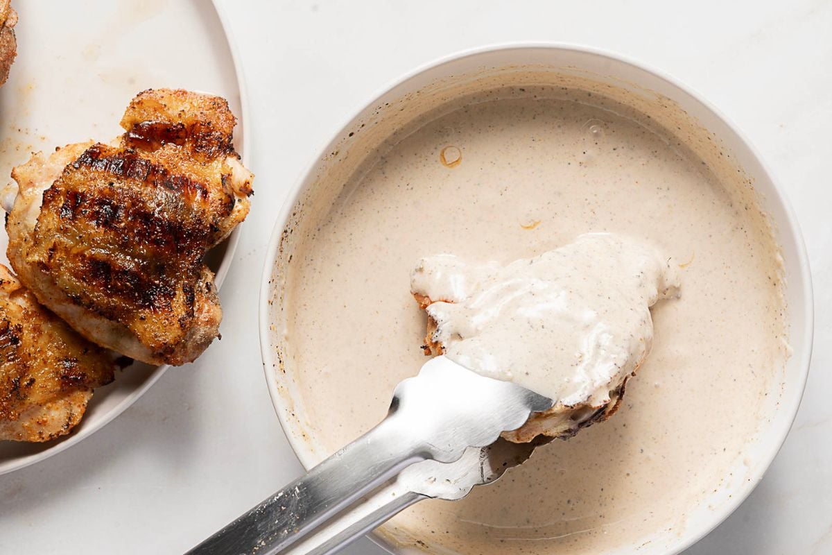 Grilled chicken is being dipped into a bowl of white sauce with metal tongs; additional grilled chicken pieces are on a plate nearby.