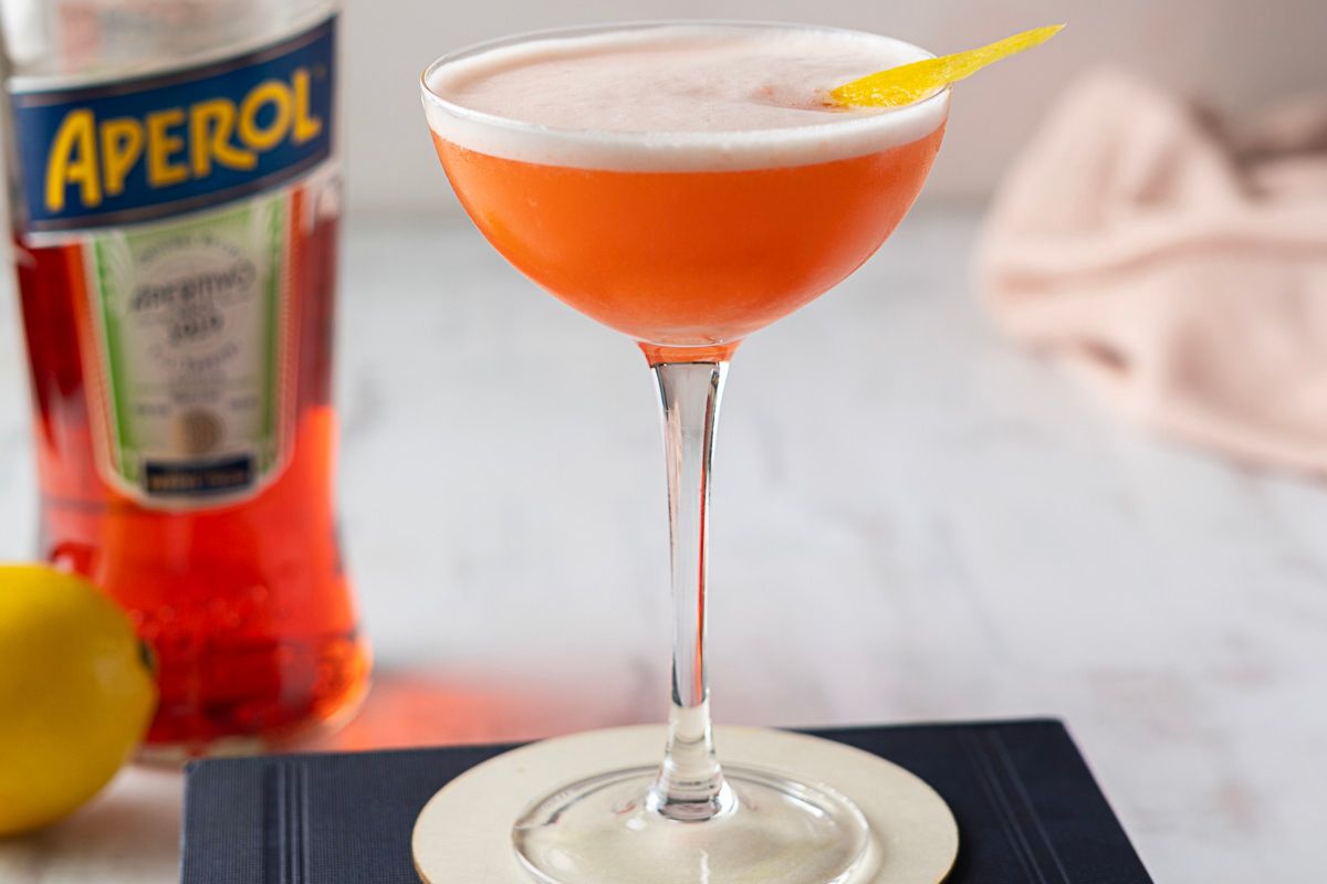 A cocktail with an orange hue and frothy top, garnished with a lemon twist, sits on a coaster; a bottle of Aperol and a lemon are in the background.