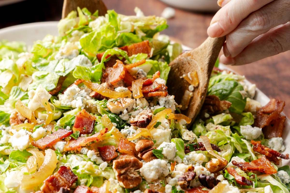 A close-up of a salad with lettuce, crumbled blue cheese, bacon pieces, walnuts, and a hand tossing it with wooden salad servers.