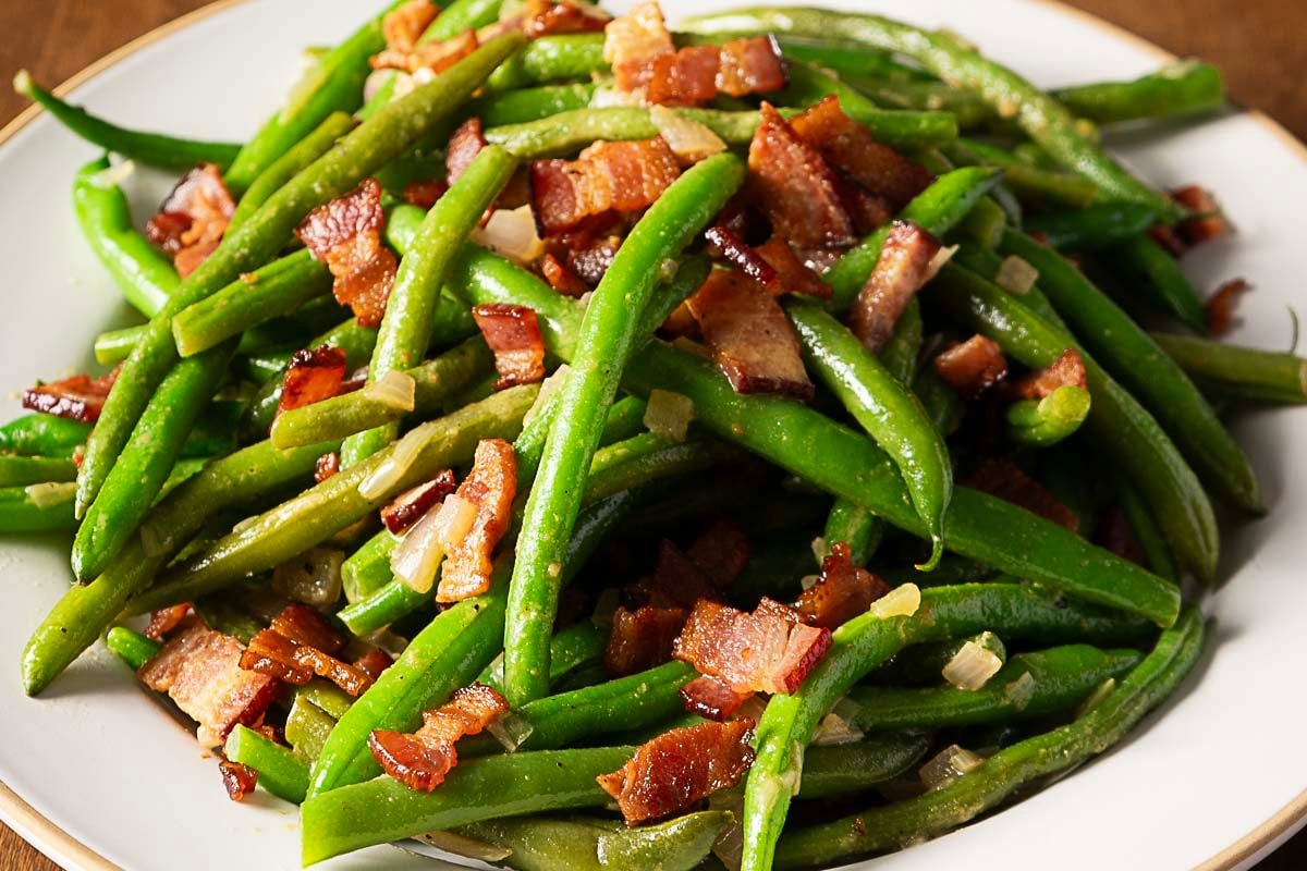 A plate of cooked green beans topped with chopped pieces of cooked bacon and small bits of onion.