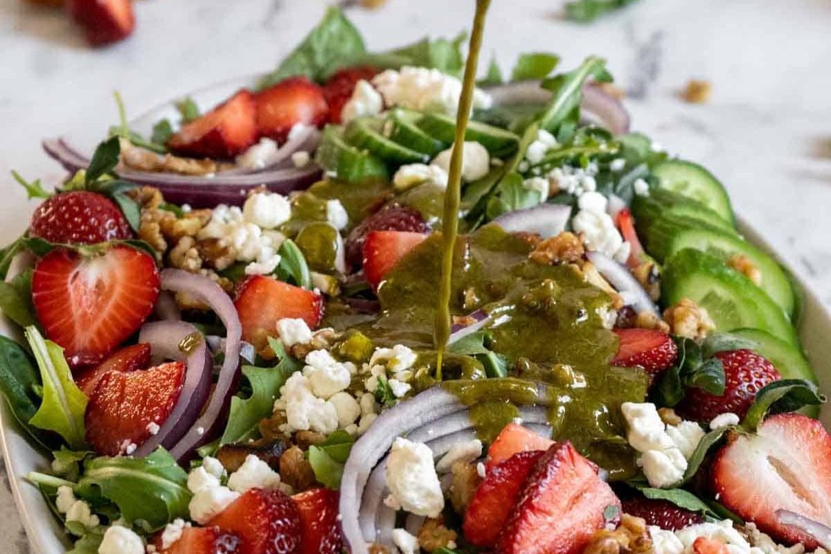 A fresh salad with sliced strawberries, cucumber, red onion, walnuts, and feta cheese being drizzled with a green dressing.