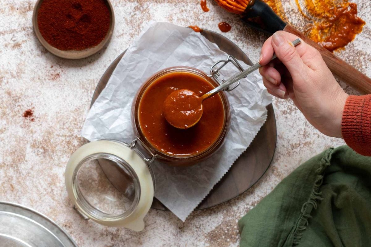 A hand holds a spoon over an open jar of reddish-brown sauce on parchment paper, with a bowl of spices and a basting brush nearby.
