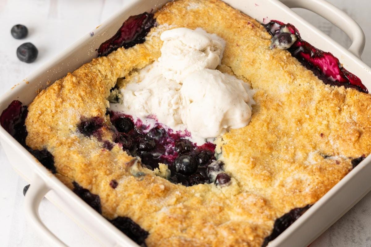 A baked blueberry cobbler topped with three scoops of vanilla ice cream in a white square baking dish.