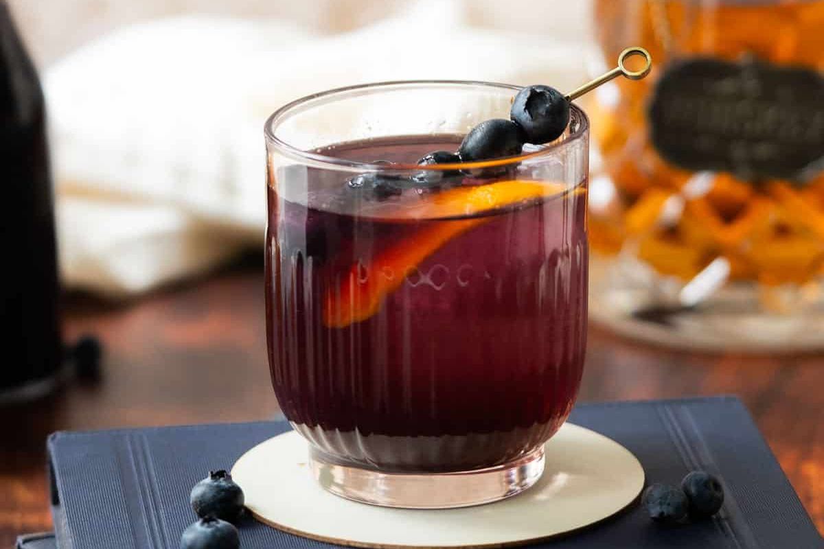 A glass of dark purple cocktail garnished with blueberries on a skewer and an orange peel, placed on a coaster with blueberries nearby.