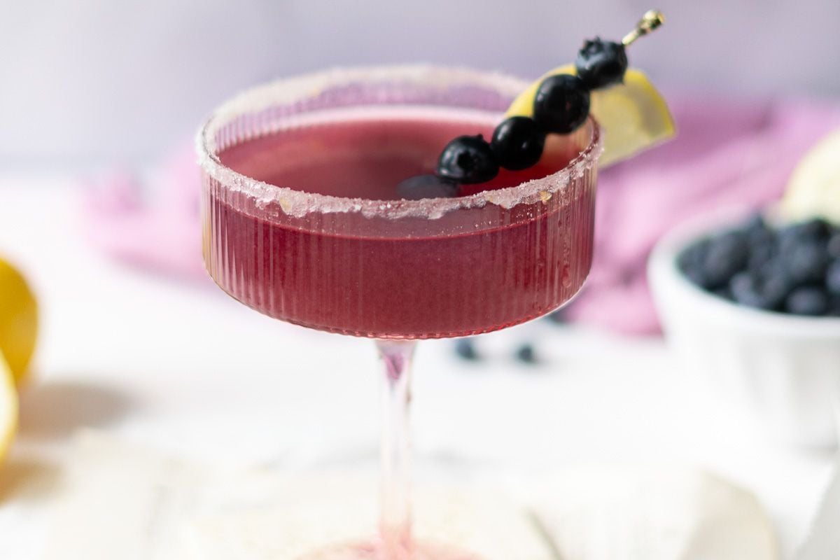 A purple cocktail in a sugar-rimmed glass, garnished with a lemon wedge and blueberries on a skewer, with blueberries and a lemon in the background.