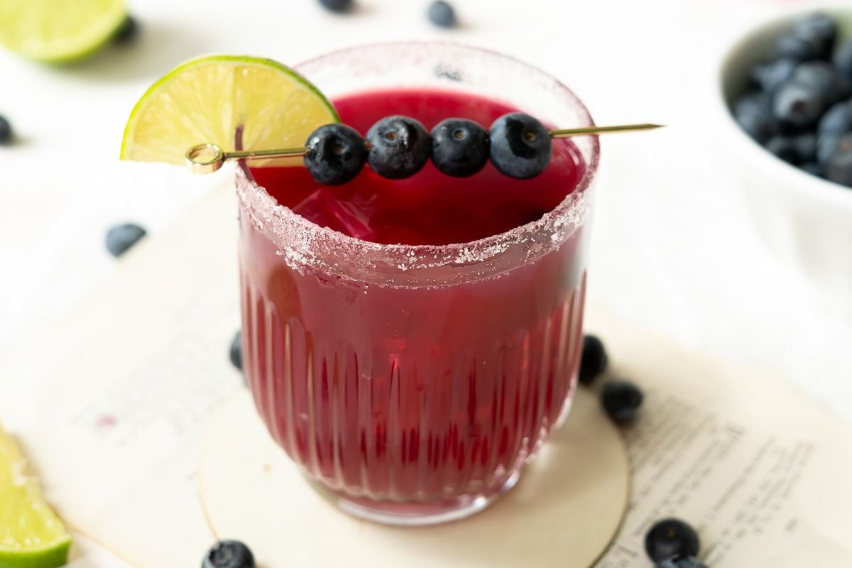 A glass of red drink with a sugared rim, garnished with a lime wedge and skewered blueberries, sits on a coaster next to scattered blueberries and a bowl of blueberries in the background.