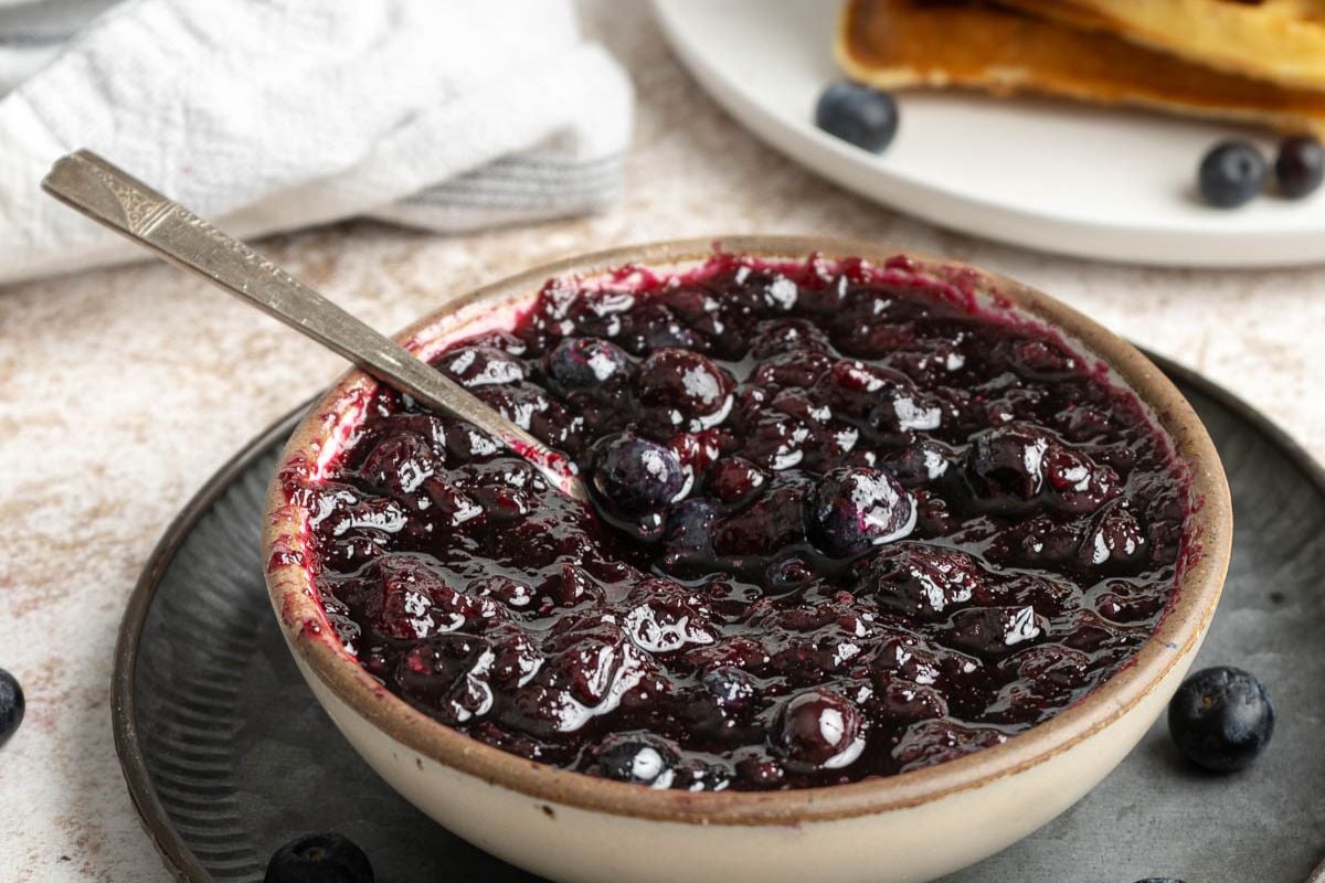 A bowl of blueberry jam with a spoon inside, placed on a dark plate with a few loose blueberries nearby.