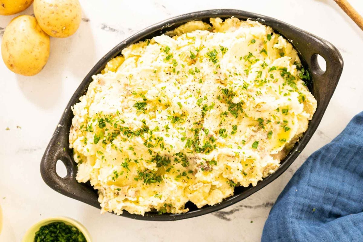 An oval baking dish filled with mashed potatoes, topped with chopped herbs and black pepper, sits on a white surface near whole potatoes and a blue cloth napkin.