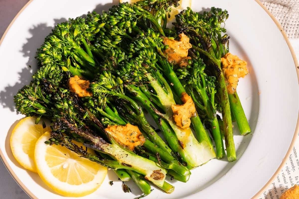 Grilled broccolini topped with dollops of orange sauce, served on a white plate with lemon slices on the side.