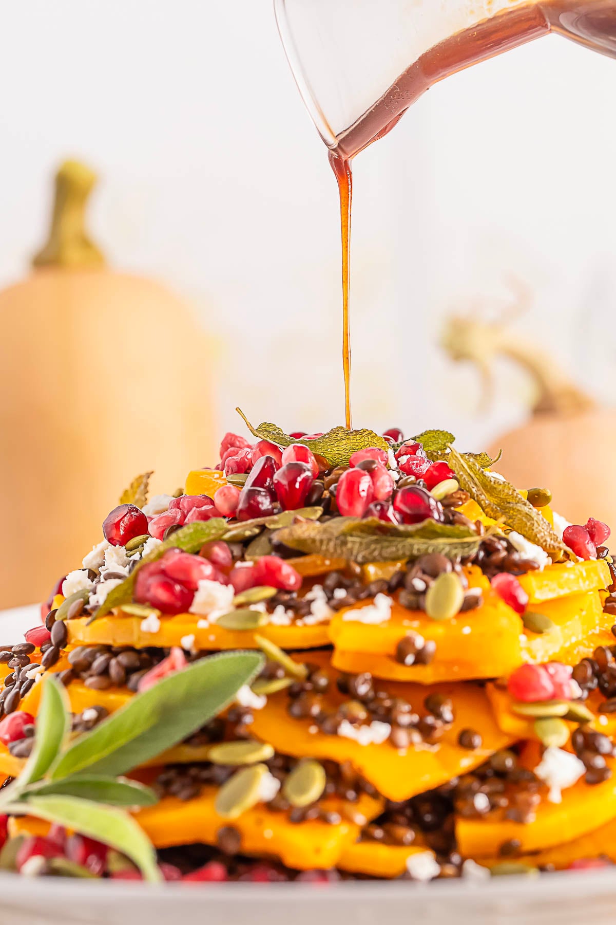 A close-up of a salad with layers of sliced squash, lentils, pomegranate seeds, pumpkin seeds, feta, and herbs, with dressing being poured on top.