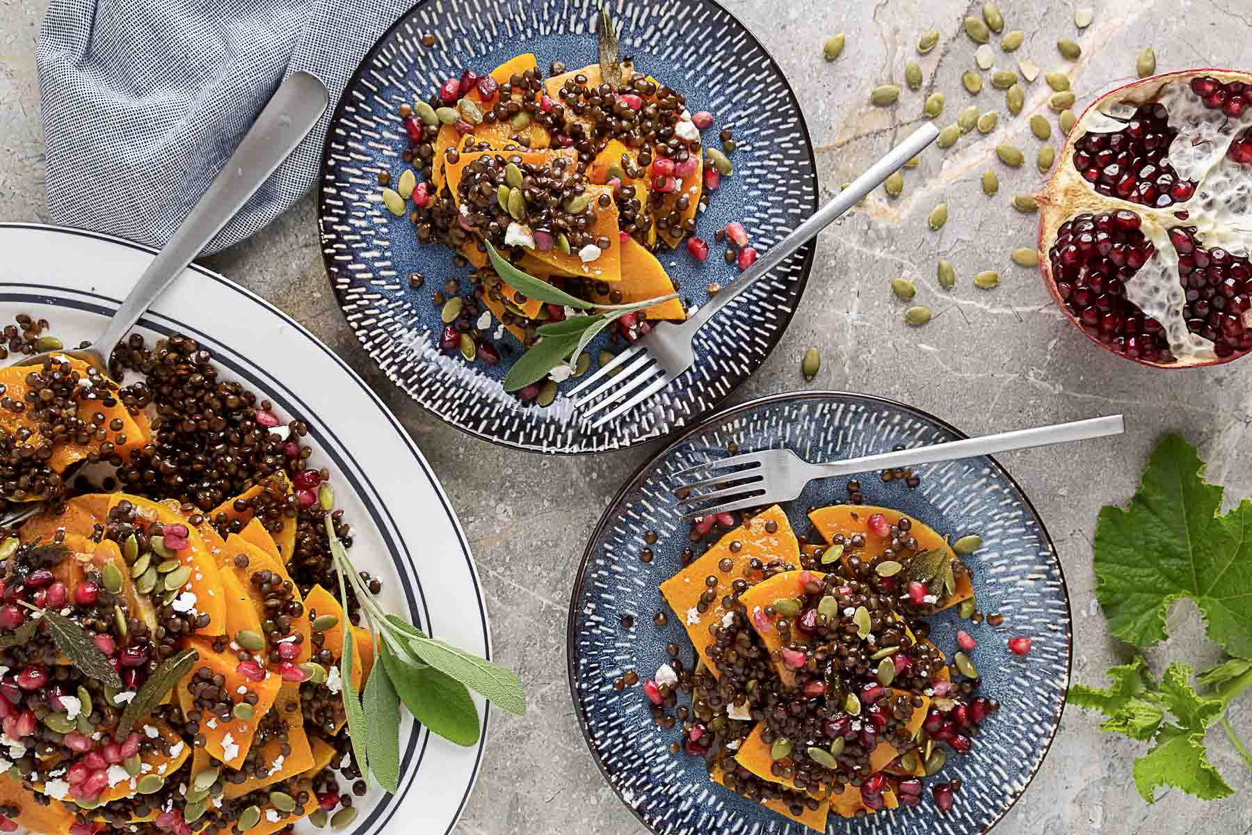 Overhead view of plates with roasted squash, lentils, pomegranate seeds, feta, and pumpkin seeds, next to a halved pomegranate and a fork.