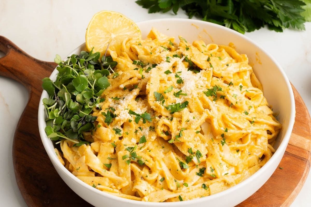 A bowl of fettuccine pasta topped with creamy sauce, grated cheese, chopped herbs, lemon wedge, and fresh greens, served on a wooden board.