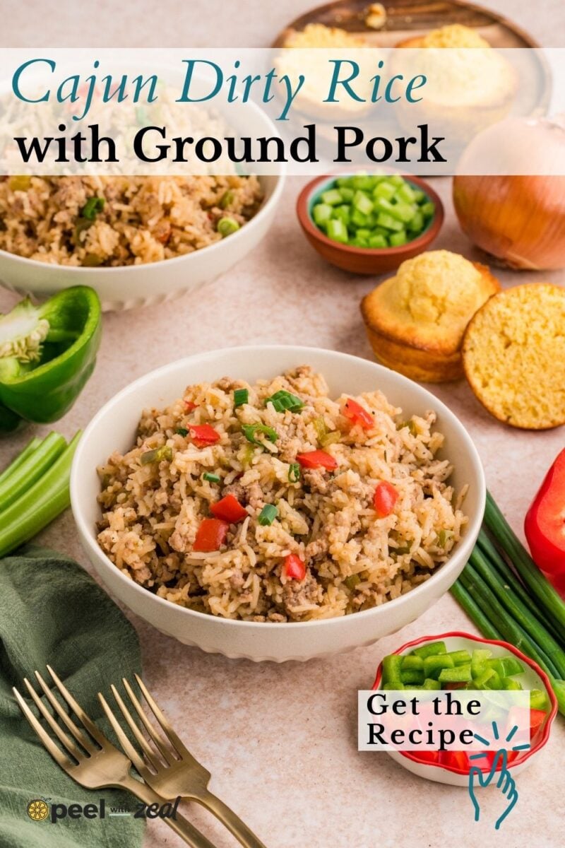 A bowl of Cajun dirty rice with ground pork, topped with diced red and green peppers, surrounded by cornbread, vegetables, and fresh ingredients on a table.