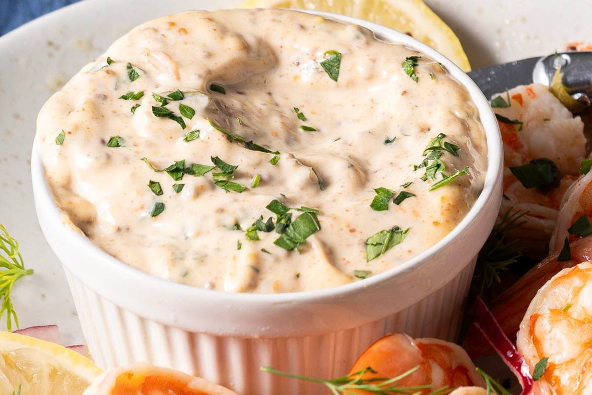 A small white ramekin filled with creamy dip, garnished with chopped herbs, sits on a plate next to shrimp and lemon wedges.