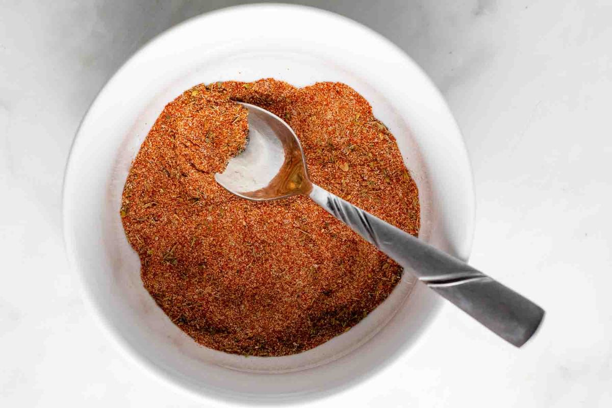 A metal spoon rests in a white bowl filled with a reddish-brown spice blend on a light surface.