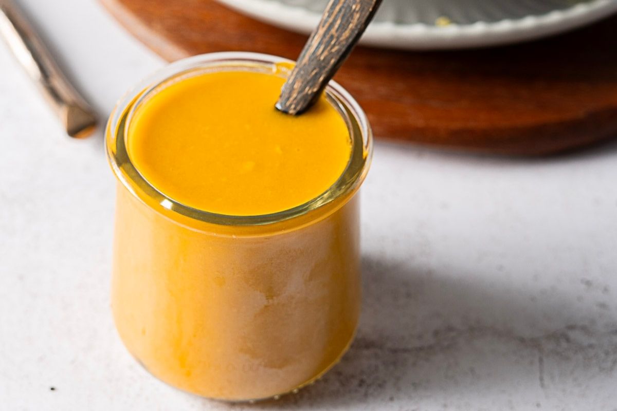 A small glass jar filled with creamy yellow sauce or dressing, with a wooden spoon inside, on a light-colored surface.