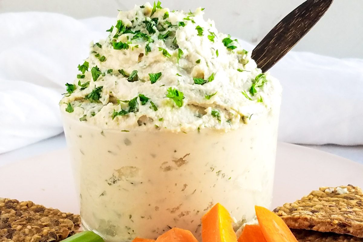 A bowl of creamy dip garnished with chopped herbs, surrounded by crackers and sliced carrots on a white plate.
