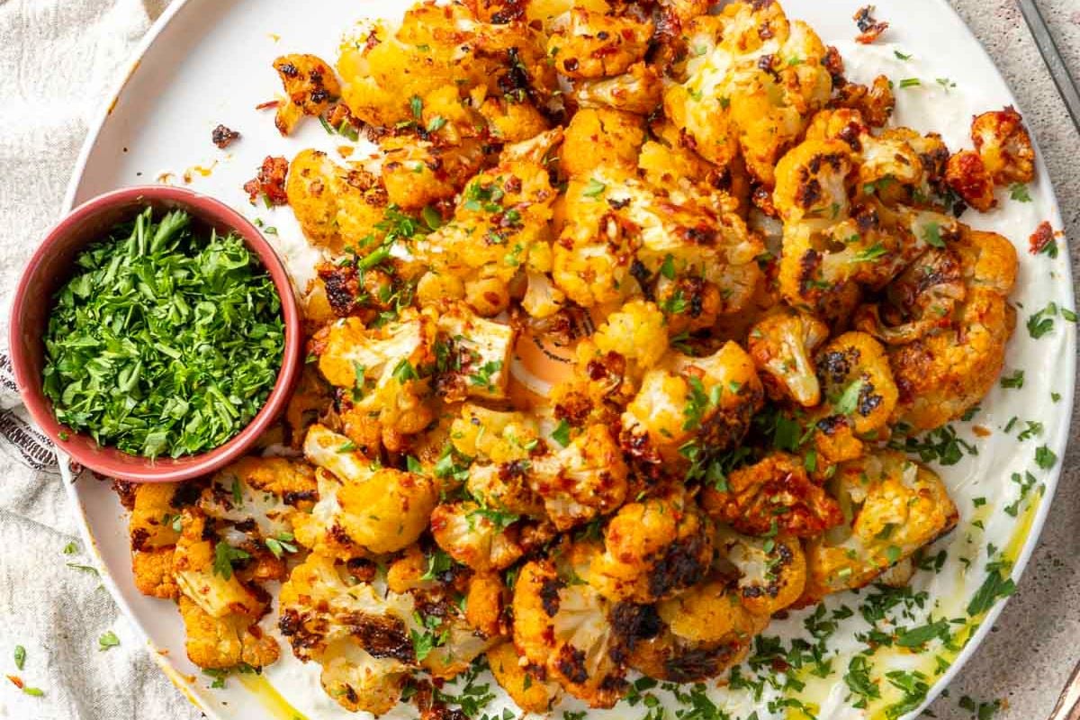 Roasted cauliflower florets garnished with chopped herbs, served on a white plate with a small bowl of fresh chopped parsley on the side.