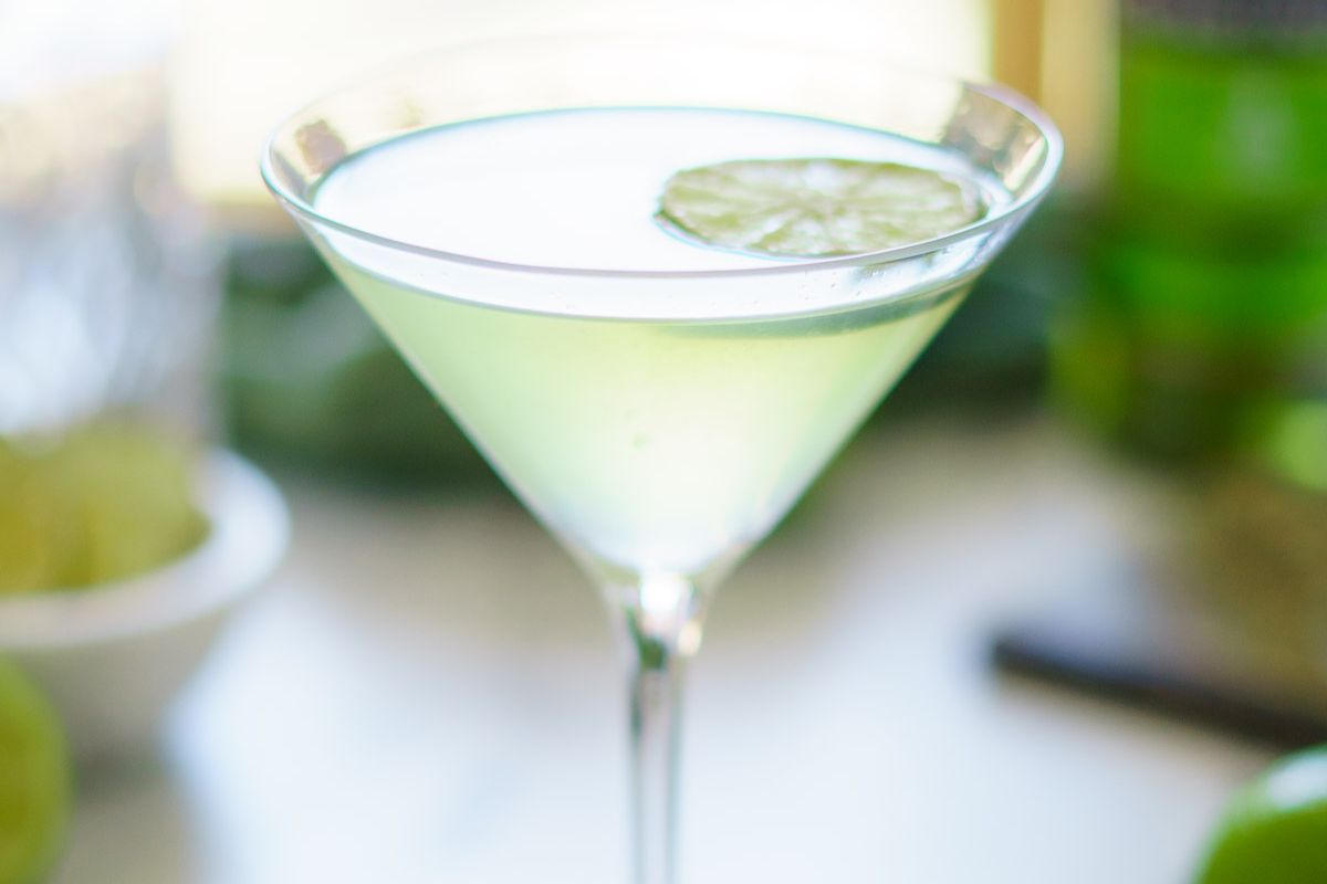 A clear martini glass filled with a pale green cocktail, garnished with a slice of lime floating on top.