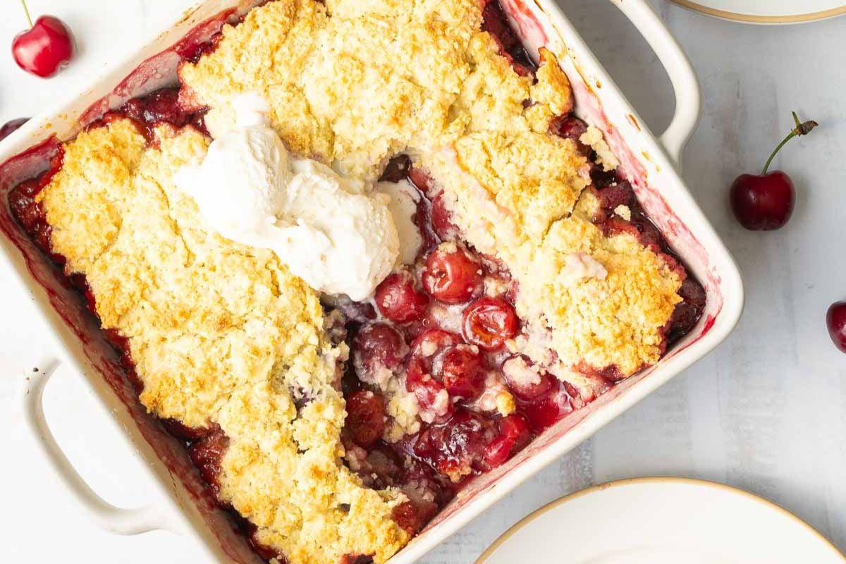 A square baking dish filled with cherry cobbler topped with golden crust and a scoop of whipped cream, with fresh cherries and plates nearby.