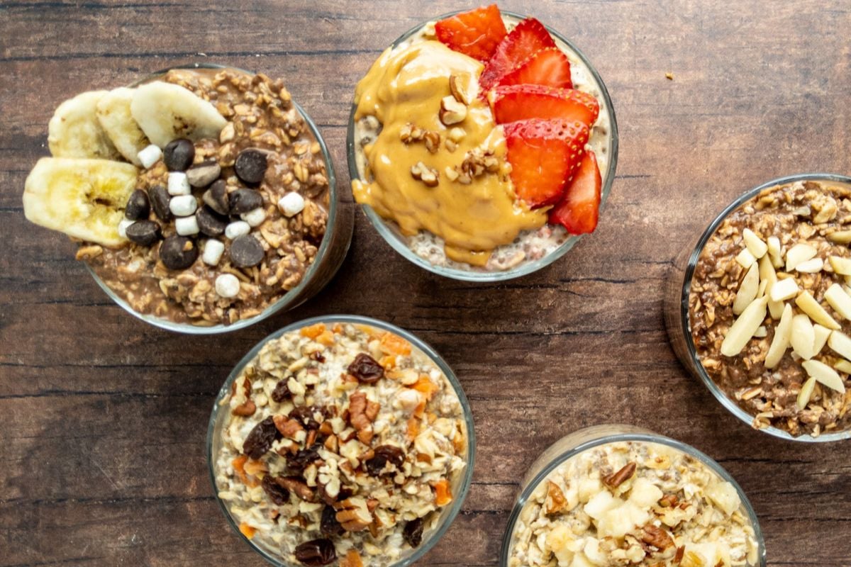 Five bowls of overnight oats with various toppings including banana slices, chocolate chips, mini marshmallows, strawberries, peanut butter, nuts, and almonds on a wooden surface.
