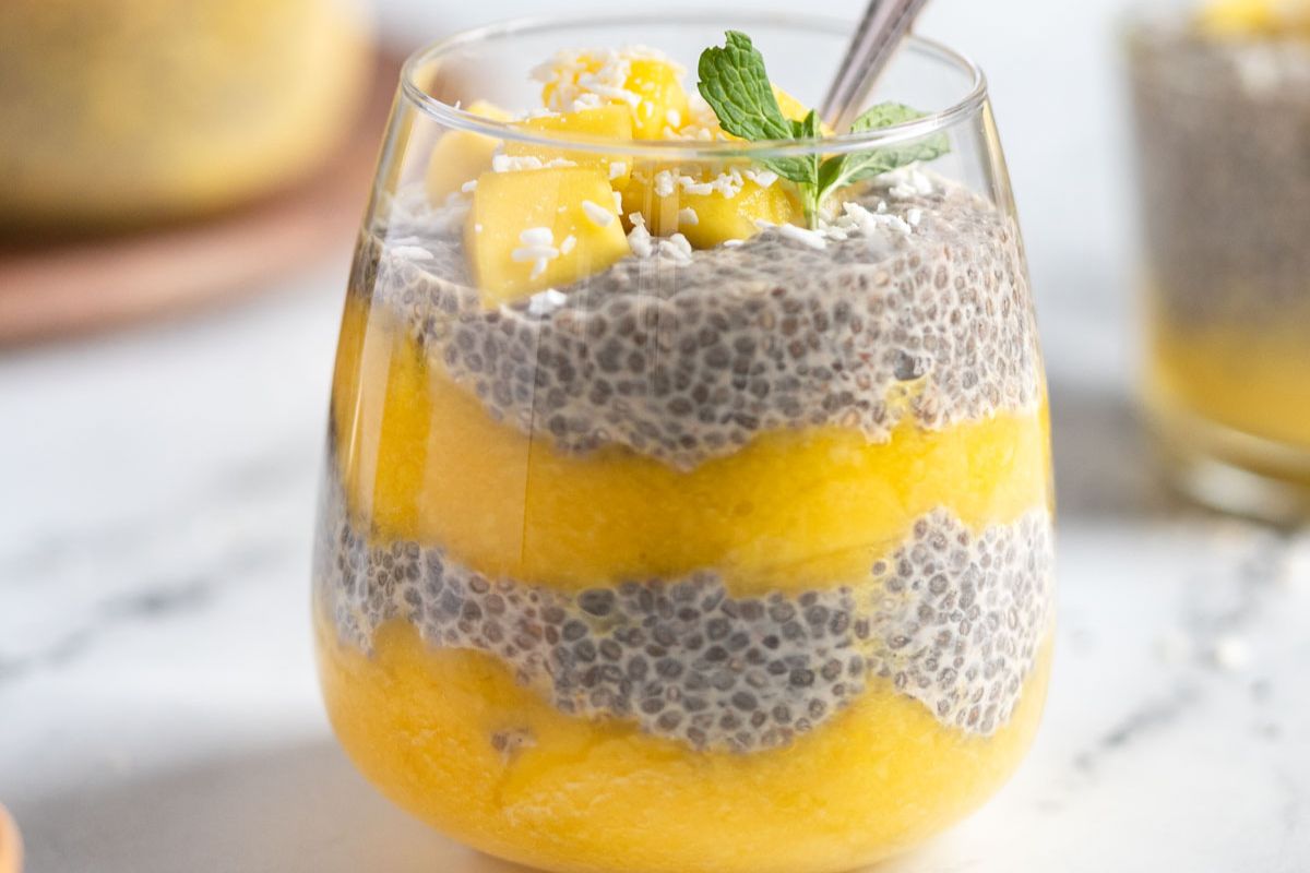A glass filled with alternating layers of mango puree and chia seed pudding, garnished with diced mango, shredded coconut, and a mint sprig.