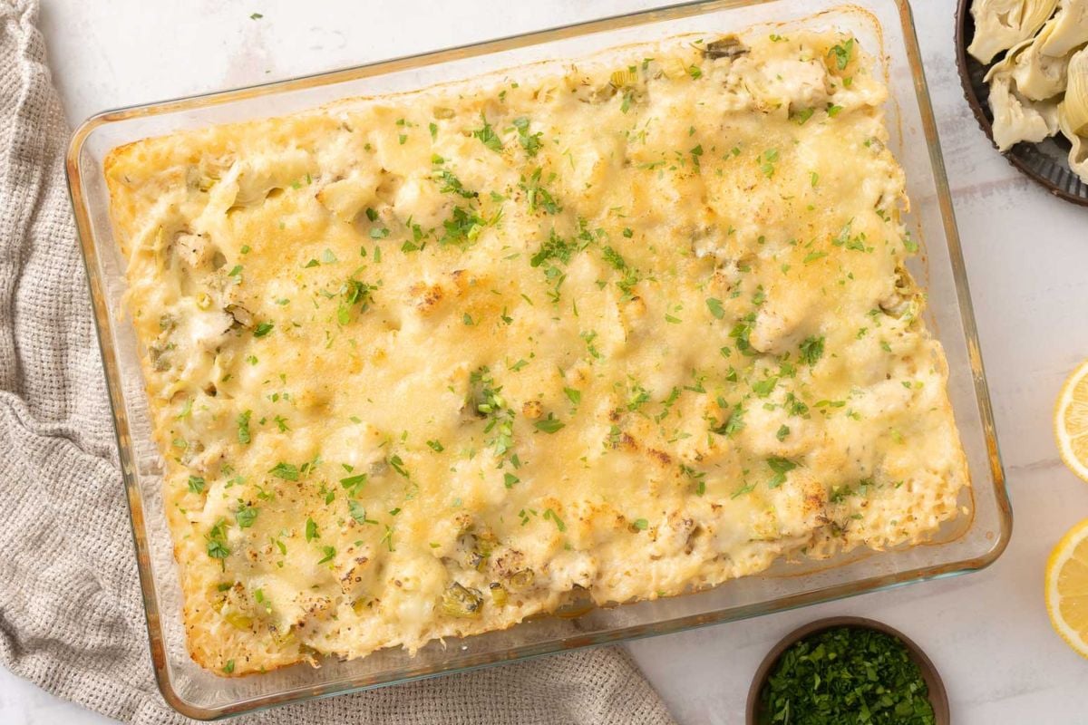 A baked casserole topped with melted cheese and chopped herbs in a glass dish, placed on a light surface next to a towel, a bowl of herbs, and lemon slices.