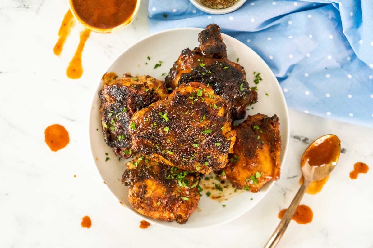 Plate of grilled chicken thighs topped with chopped herbs, with a spoon, sauce stains, and a blue polka dot napkin nearby on a white surface.