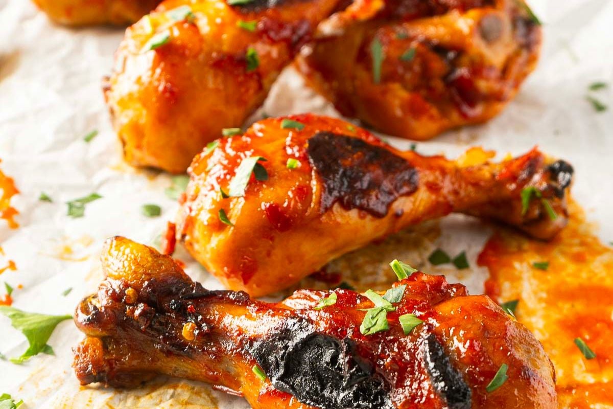 Close-up of baked chicken drumsticks coated in a reddish glaze, garnished with chopped herbs, on parchment paper.