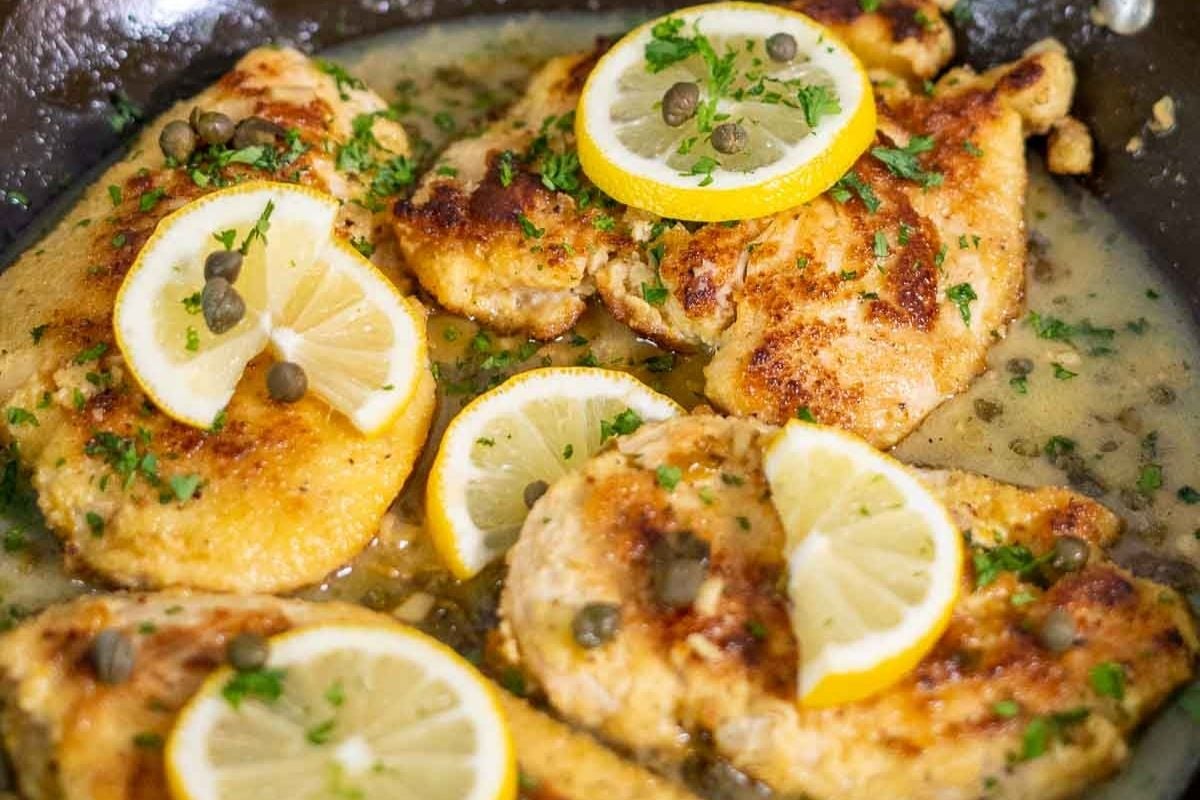 Cooked chicken breasts in a skillet, topped with lemon slices, capers, and chopped parsley in a light sauce.