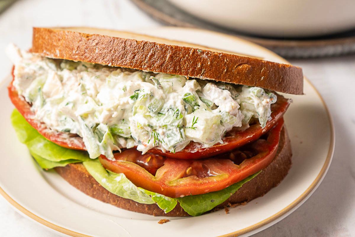 A chicken salad sandwich with lettuce and tomato on brown bread, served on a white plate.