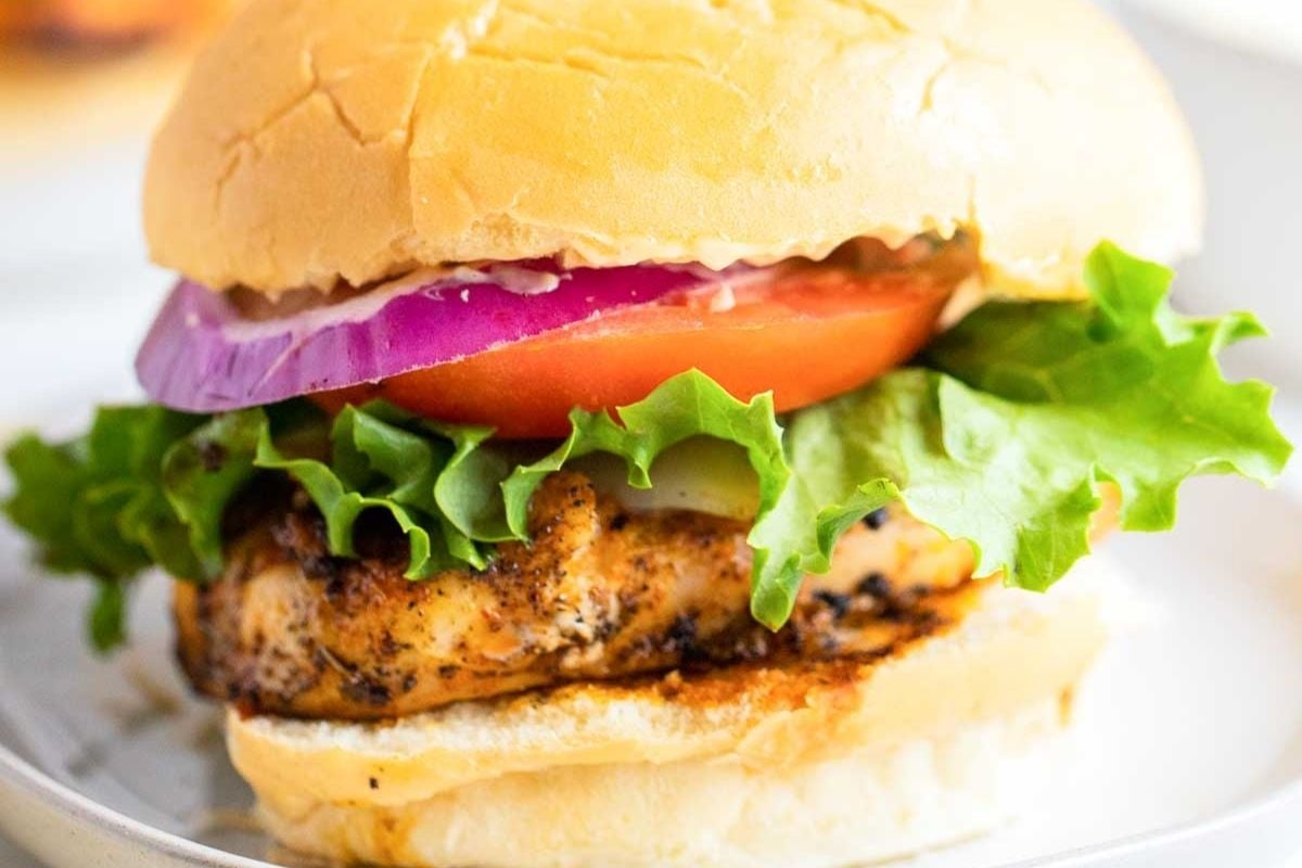 A grilled chicken sandwich with lettuce, tomato, red onion, and mayonnaise on a bun, served on a white plate.
