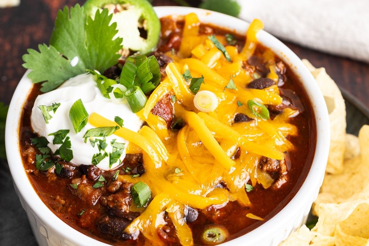 A bowl of chili topped with shredded cheddar cheese, sour cream, sliced green onions, cilantro, and jalapeño, with tortilla chips on the side.