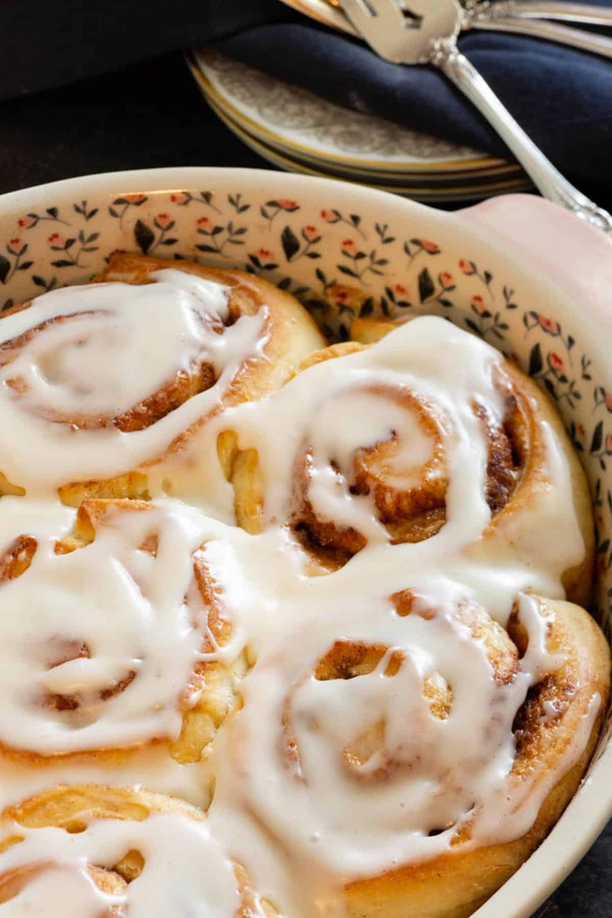 A close-up of iced cinnamon rolls in a decorative baking dish, with a serving spoon and stacked plates in the background.