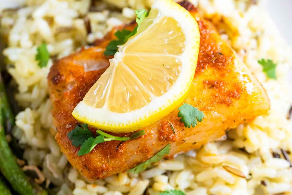 A cooked salmon fillet topped with a lemon slice and cilantro sits on a bed of rice with green beans on the side.