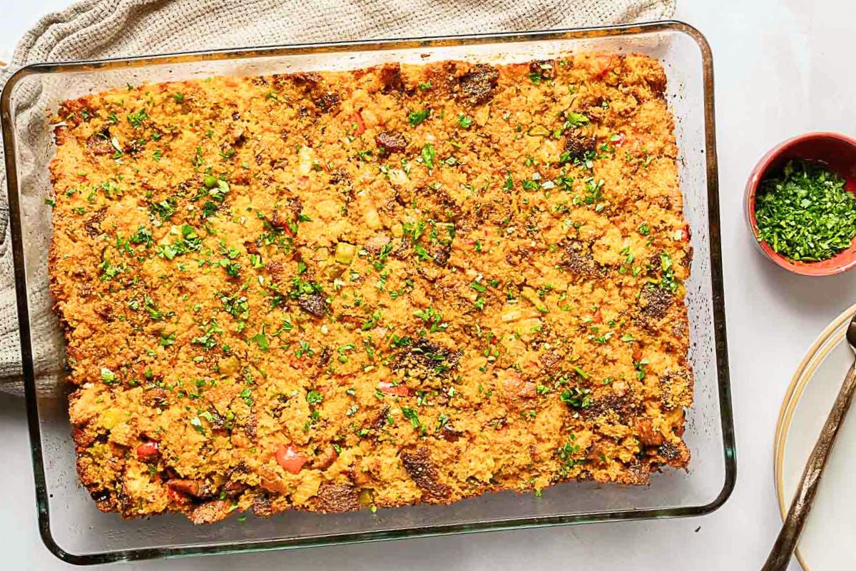 A glass baking dish filled with baked cornbread stuffing topped with chopped herbs, placed on a neutral cloth with a bowl of herbs and a serving spoon nearby.