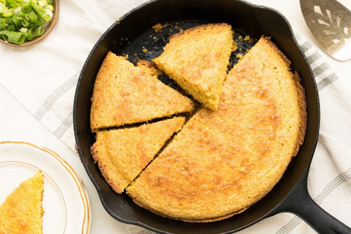 A cast iron skillet with golden cornbread, partially sliced, sits on a white cloth next to a plate and a bowl of chopped green onions.
