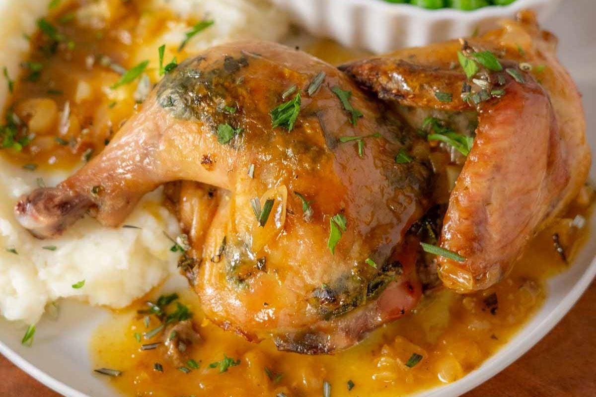 Roast chicken with herb sauce served on mashed potatoes, garnished with chopped parsley, with a side of green peas in the background.