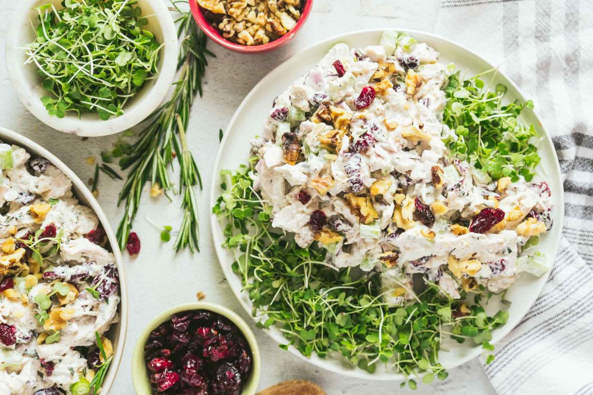 A plate of chicken salad with microgreens, walnuts, and dried cranberries—perfect for those seeking easy chicken recipes—surrounded by small bowls of walnuts, cranberries, and microgreens on a white surface.