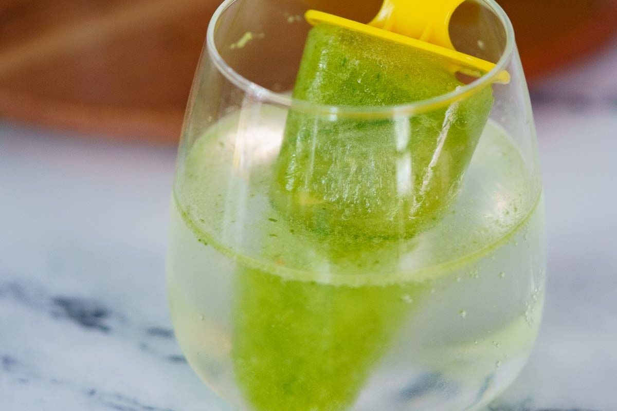 A green popsicle on a yellow stick is placed in a clear glass of a transparent, bubbly beverage on a white surface.