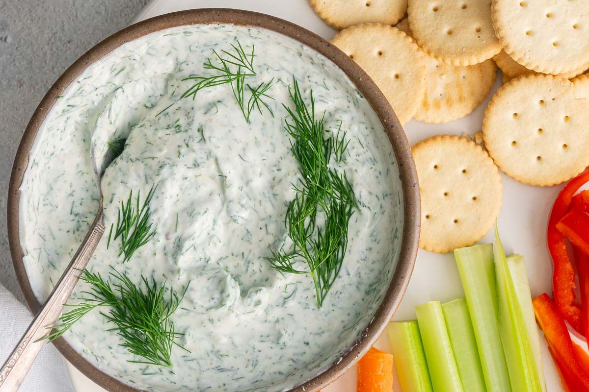 A bowl of creamy dill dip garnished with fresh dill, surrounded by round crackers, celery sticks, carrot sticks, and red bell pepper slices.