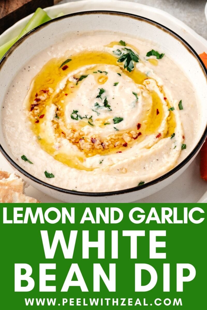 A bowl of creamy white bean dip garnished with olive oil, herbs, and red pepper flakes, surrounded by fresh veggie sticks and pita bread.