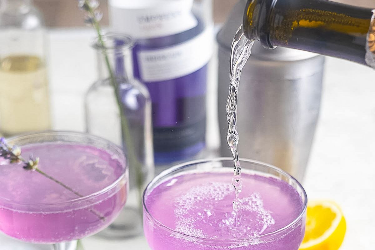 A sparkling liquid is being poured into a coupe glass with a purple cocktail. Other bottles, a shaker, and a lemon half are on the table in the background.