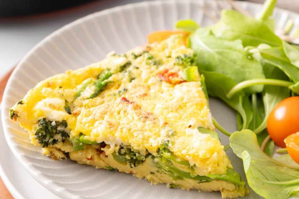A slice of vegetable frittata with broccoli and herbs is served on a plate next to fresh leafy greens and cherry tomatoes.