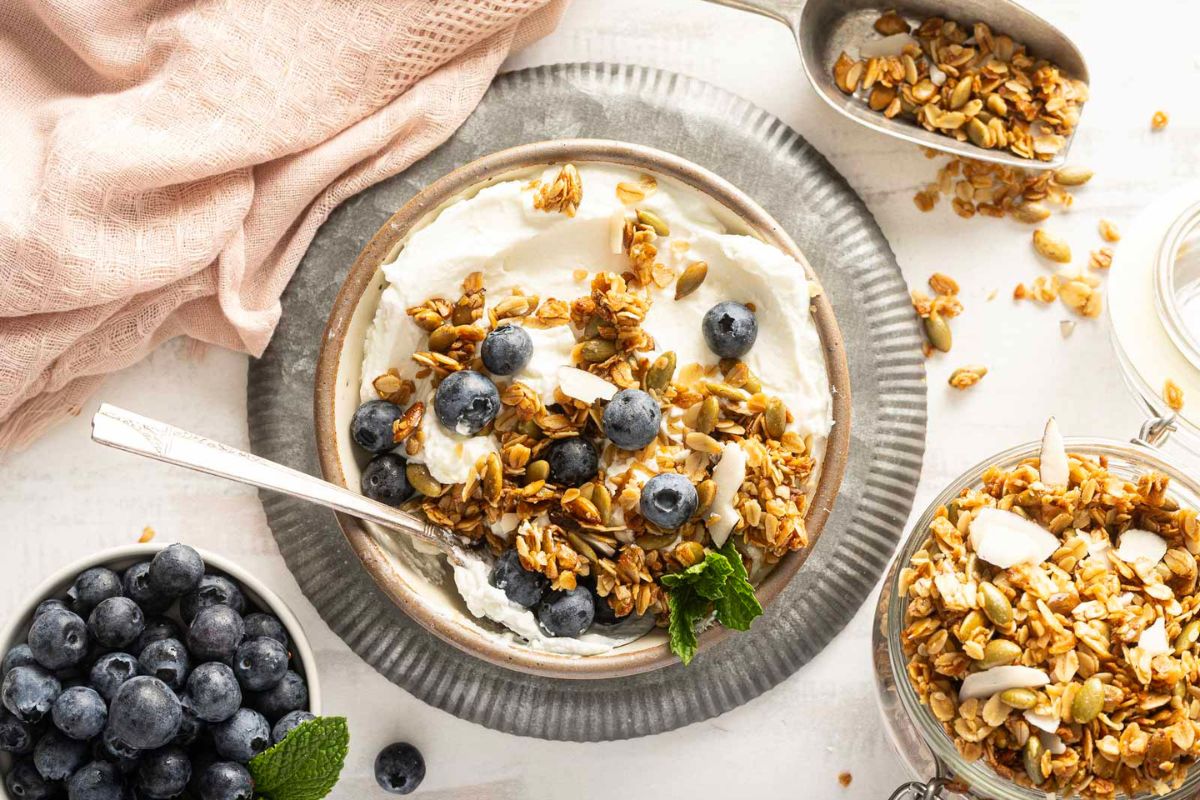 A bowl of yogurt topped with granola and blueberries sits on a metal plate with a spoon, surrounded by a bowl of blueberries, a jar of granola, and a pink cloth.